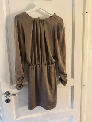 Beige kort klänning med knytband - Säljer en beige kort klänning med långa ärmar och ett snyggt knytband i midjan. Klänningen har en avslappnad passform och är tillverkad i ett mjukt material som ger ett fint fall. Perfekt för dig som gillar enkel men stilren design.