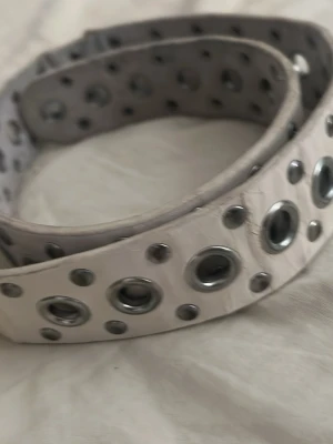 Vitt bälte med silverhål y2k vintage - Snyggt vitt bälte med massor av silverfärgade metallnitar längs hela bandet. Bältet har ett edgy och trendigt uttryck, perfekt för att lyfta vilken outfit som helst. Gjort i syntetmaterial och har rundade hål med metallringar. Slitet
