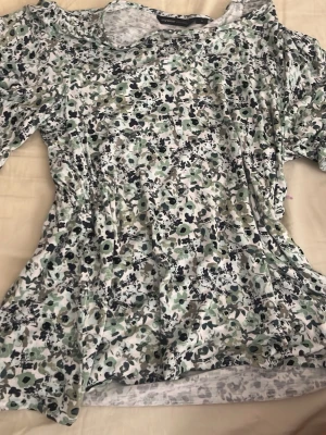 Blommig topp med cut out-detaljer - Snygg topp med blommigt mönster i vitt, svart och grönt. Modellen har cut out-detaljer vid axlarna och är tillverkad i ett mjukt, stretchigt material. Perfekt för dig som gillar en feminin och trendig stil.