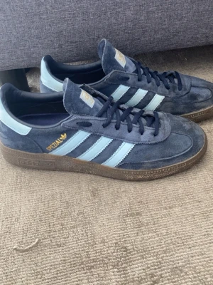 Adidas Spezial blå sneakers mocka - Säljer ett par Adidas Spezial sneakers i blå mocka med ljusblåa ränder och detaljer. Skorna har klassisk låg profil, bruna gummisulor och guldfärgad Spezial-logga på sidan. Perfekta för dig som gillar retrostil och streetwear.