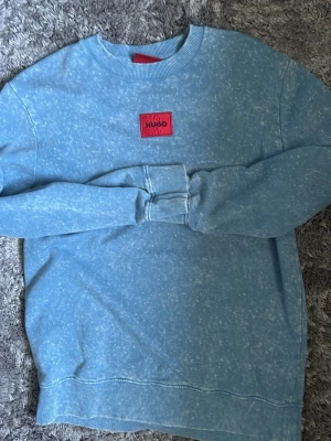 Ljusblå sweatshirt från Hugo - Säljer en ljusblå sweatshirt från Hugo med en röd patch med logga på bröstet. Tröjan har rund halsringning, långa ärmar och ribbade muddar vid ärmslut och nederkant. Perfekt för en avslappnad och stilren look.