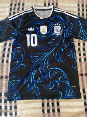 Argentina Messi tröja svart/blå Adidas - Snygg svart fotbollströja från Adidas med blått mönster, nummer 10 och Messi på ryggen. Framsidan har FIFA-märke och AFA-logga. Korta ärmar och klassiska vita ränder på axlarna. Perfekt för dig som älskar fotboll och vill sticka ut på planen. Helt ny med tagg. Priset är ej hugget i sten!