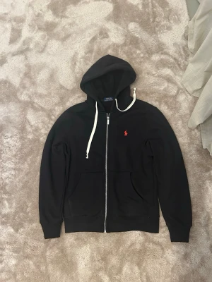 Svart hoodie från Polo Ralph Lauren - Svart hoodie med dragkedja från Polo Ralph Lauren. Klassisk design med vit dragsko i huvan och röd broderad logga på bröstet. Mjuk bomullskänsla och två fickor framtill. Perfekt för en avslappnad och stilren look. Kan gå ner i pris vid snabb affär. Snörerna är knutna för att de var för långa går att ta bort det