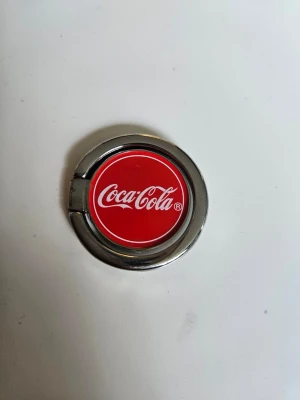 Coca-Cola mobilring i metall - Säljer en snygg mobilring med klassisk Coca-Cola-logga. Ringen är rund, har en röd framsida med vit text och en silvrig metallram. Perfekt för bättre grepp på mobilen eller som stativ. Självhäftande baksida för enkel montering.