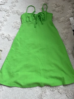 Grön sommarklänning med snörning - Säljer en grön klänning med smala axelband och snörning framtill. Klänningen har en lätt utställd passform och är tillverkad i ett strukturerat tyg som känns luftigt. Perfekt för varma dagar och enkel att styla med sneakers eller sandaler.