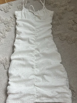 Vit kort klänning med snörning - Säljer en vit kort klänning med smala axelband och snörning fram. Klänningen har ett broderat hålmönster och är figurnära med rynkade detaljer längs mitten. Perfekt för sommarens alla tillfällen!