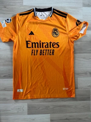 Real Madrid orange Adidas - Snygg orange Real Madrid  från Adidas med svart klubbmärke och sponsortryck på bröstet. Tröjan har svarta detaljer vid axlar och krage samt Champions League-märke på ärmen. Tillverkad i lätt och ventilerande material.
