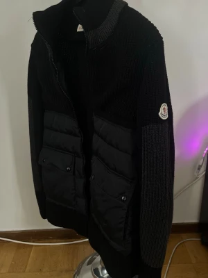 Moncler Cardigan - Snygg svart cardiganjacka från Moncler med stickade ärmar och quiltade partier framtill. Jackan har dragkedja, två stora fickor med lock och Moncler-logga på ärmen. Perfekt för dig som vill ha en stilren och exklusiv look.