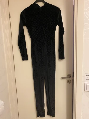 Byxdress - Svart byxdress med mönster/bokatäver på. Dragkedja fram, ner till ungefär naveln (se bild nummer 3) Materialet på känns lite som sammet. Bälte följer med (se bild nummer 6). Står storlek S på, men tycker att det passar en XS bättre. Aldrig använd!