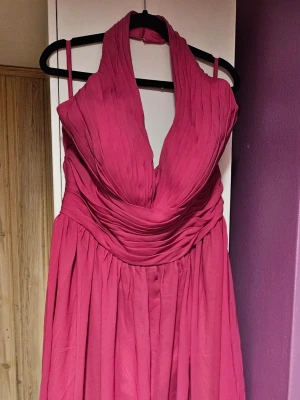 Rosa långklänning från Hebeos - Säljer en helt ny snygg rosa långklänning från Hebeos med djup v-ringning och halterneck. Klänningen har plisserad överdel och ett svepande, elegant fall. Perfekt för dig som vill sticka ut med en färgstark look. 
