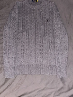 Grå kabelstickad tröja Polo Ralph Lauren - Kabelstickad grå tröja från Polo Ralph Lauren med rund halsringning och ribbade muddar. Klassisk broderad logga på bröstet. Perfekt för lager på lager och en stilren look. Materialet känns mjukt och håller värmen.