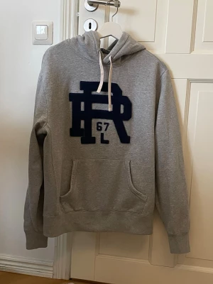 ”Rare” hoodie Polo Ralph Lauren - Snygg grå hoodie från Polo Ralph Lauren med stor mörkblå PR 67-logga på bröstet. Klassisk känguruficka framtill och vita dragsnören i huvan. Tillverkad i mjuk bomullsblandning, perfekt för en avslappnad streetwear-look.