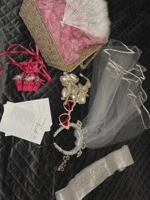 Brudaccessoarer till möhippa - Korg med vit slöja, diadem med texten 'BRIDE', vitt band med “BRIDE” glittertext, hjärtformade glasögon, beiga ballonger, rosa visselpipor och spelkort för möhippa.