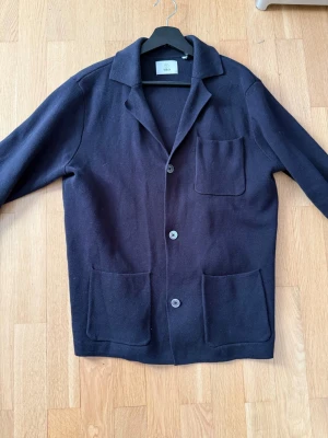 Mörkblå cardigan från bläck - Stilren mörkblå cardigan från bläck med tre stora framfickor och klassisk krage. Jackan har tre knappar framtill och är tillverkad i ett mjukt material som ger en avslappnad men ändå uppklädd look. Perfekt för lager på lager under kyligare dagar.