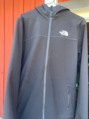 Svart jacka från The North Face - Svart hoodie med hel dragkedja och huva från The North Face. Jackan har två sidofickor med dragkedja och vit logotyp på bröstet och ryggen. Materialet är slätt och funktionellt, perfekt för att hålla dig varm och skyddad mot vind.