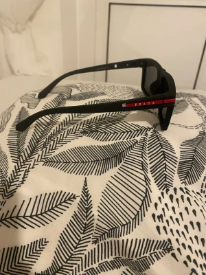 Svarta solglasögon från Prada - Snygga svarta solglasögon från Prada med en röd detalj och logga på skalmen. Modellen har en klassisk, rak form och är tillverkad i ett tåligt syntetmaterial. Perfekta för dig som vill ha en stilren och exklusiv look. Fick den som en studentpresent men kom aldrig till användning 