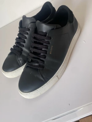 Svarta sneakers från Axel Arigato - Stilrena svarta sneakers från Axel Arigato med svart läder, svarta skosnören och vit sula. Diskret guldtext på sidan och klassisk låg modell som passar till det mesta. Ovandelen är i slätt läder och sulan är i gummi.