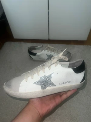Golden Goose vita sneakers med glitterstjärna - Säljer ett par vita Golden Goose sneakers med silverglittrig stjärna på sidan och svart detalj vid hälen. Skorna har beige snörning och grå detaljer vid tån. Klassisk låg modell i läder som passar perfekt till streetwear och casual outfits. OBS: Står 44 men passar 43 