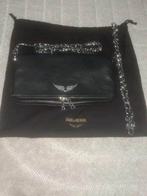 Svart axelväska Zadig & Voltaire - Knappt använt Zadig & Voltaire väska, köpte den på jackie skriv om ni vill ha flera bilder💓 Cool svart axelväska i läder från Zadig & Voltaire med silverfärgad kedjerem och ikonisk vinge i metall framtill. Väskan har dragkedja och logga på framsidan, perfekt för dig som gillar en edgy och stilren look. 