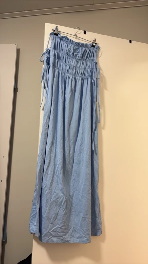 Ljusblå maxiklänning med smock - Ljusblå maxiklänning med smock upptill och knytband på axlarna. Klänningen har en avslappnad passform och är gjord i ett mjukt, tunt tyg som faller snyggt. Perfekt för en somrig look med sin enkla men trendiga design.