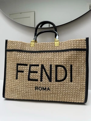 Fendi inspirerad shoppingväska i strå - Säljer en snygg Fendi inspirerad shoppingväska i naturfärgat flätat strå med svarta detaljer och stora FENDI ROMA-bokstäver framtill. Väskan har svarta handtag och guldfärgade metalldetaljer med Fendi-logga. Perfekt för dig som vill ha en trendig och rymlig väska.