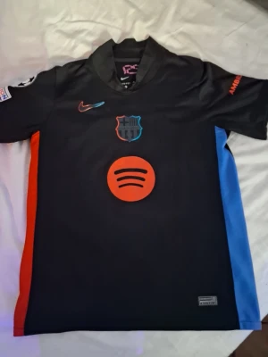 FC Barcelona svart matchtröja Nike - Snygg svart FC Barcelona matchtröja från Nike med röda och blå detaljer. Tröjan har Spotify-logga på bröstet, klubbmärke och Nike Swoosh. På ryggen står det 'Lamine Yamal' och nummer 19 i rött. Perfekt för dig som älskar fotboll och Barca. 
