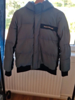 Grå och svart pufferjacka Calvin Klein - Snygg pufferjacka från Calvin Klein i grått och svart med huva och svart logotypdetalj på bröstet. Jackan har svarta muddar och nederkant, samt en quiltad design som håller värmen. Perfekt för kalla dagar och riktigt stilren look.