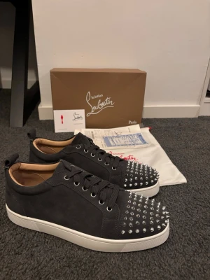 Christian Louboutin svarta spikes sneakers - Svarta sneakers i mocka från Christian Louboutin med silverfärgade nitar på tån och klassisk röd sula. Lågt skaft, svarta skosnören och vit yttersula. Perfekta för dig som vill sticka ut med en edgy och lyxig stil.