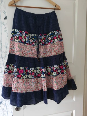 Blommig långkjol med snörning - Säljer en långkjol med mörkblå botten och färgglada blommiga partier. Kjolen har snörning i midjan. Perfekt för dig som gillar boho-stil och vill ha något unikt till sommaren.