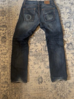 Nudie jeans  - Säljer ett par klassiska blå jeans med raka ben och snygga slitningar på både fram- och baksida. Jeansen har fem fickor och kontrastsömmar. Perfekta för en avslappnad och trendig look. Storleken är 30/32 måtten är 40cm i midjan 107cm i längden och 19cm i benöppningsmåttet 