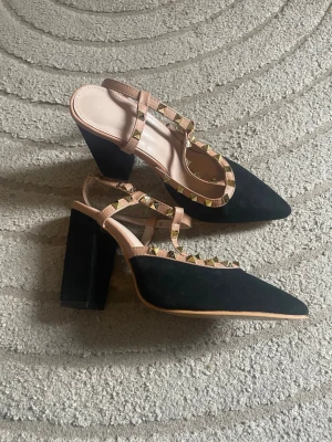 Svarta pumps med nitar och blockklack - Säljer ett par svarta pumps med bred blockklack och spetsig tå. Skorna har nude-färgade remmar dekorerade med guldfärgade nitar för en edgy look. Materialet ser ut att vara mockaimitation och syntet. Perfekta för dig som vill sticka ut lite extra.