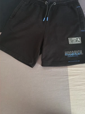 Svarta shorts från Hoodrich med snörning - Svarta shorts från Hoodrich med blå detaljer och snörning i midjan. Shortsens framsida har en patch med logotyp och text, samt fickor på sidorna. Materialet är mjukt och troligtvis bomull eller polyester, perfekt för en avslappnad stil.