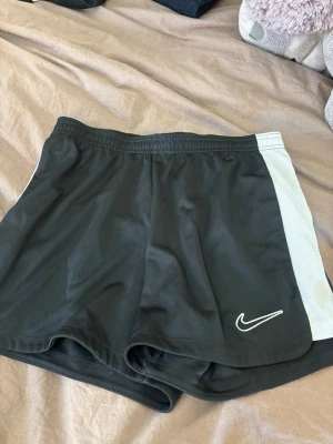 Svarta Dri-FIT shorts från Nike - Säljer ett par svarta sportiga shorts från Nike med vita sidopaneler och klassisk swoosh-logga framtill. Tillverkade i lätt och snabbtorkande Dri-FIT material som passar perfekt för träning eller varma dagar.