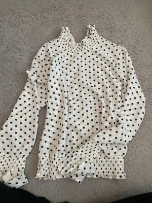 Vit prickig blus - Vit blus med svarta prickar och smock vid hals, ärmslut och midja. Långärmad modell med lätt volang vid ärmslut och hög hals. 