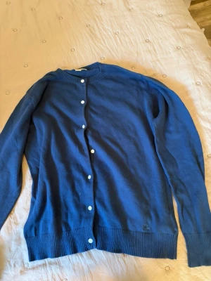 Blå kofta från Burberry - Snygg blå kofta från Burberry med rund halsringning och vita knappar framtill. Tillverkad i mjukt stickat material och har ribbade muddar vid ärmslut och nederkant. Perfekt för att addera en klassisk touch till din outfit.