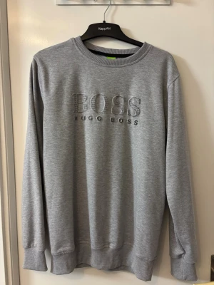 Grå sweatshirt från Hugo Boss - Snygg grå sweatshirt från Hugo Boss med broderad logga och texten 'BOSS' framtill. Tröjan har rund halsringning, ribbade muddar vid ärmslut och nederkant samt en klassisk relaxed fit. Perfekt för en clean och stilren look.