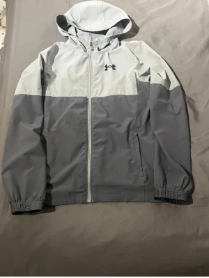 Grå vindjacka från Under Armour - Snygg tvåtonad grå vindjacka från Under Armour med huva och dragkedja framtill. Jackan har logga på bröstet och elastiska muddar med tryckt text. Perfekt för blåsiga dagar och sportiga outfits. Tillverkad i lätt, vindtätt material.