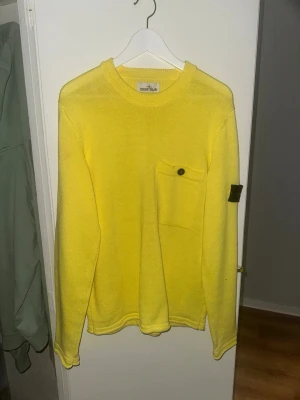 Gul stickad tröja från Stone Island - Snygg gul stickad tröja från Stone Island med rund halsringning och en bröstficka med knapp. Tröjan har lång ärm och klassisk Stone Island-patch på vänster ärm. Perfekt för dig som vill sticka ut med färg och stil.