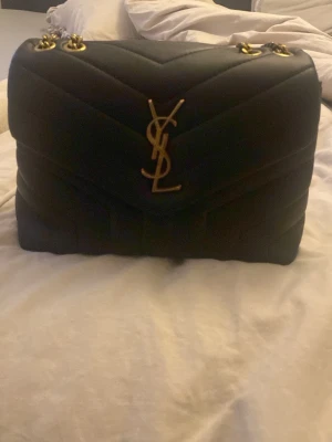 Svart YSL axelväska i läder - Säljer en svart axelväska som är köpt på Plick säljaren sa inte om den var äkta men antar att den inte är det eftersom av alla defekter den fått som rostning och skinnet på bottnen men man ser ingen av det egentligen vilket är bra