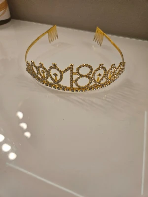 Guldigt diadem med siffran 18 - Snyggt diadem i guldfärg med siffran 18 framtill, dekorerat med glittrande strass. Perfekt accessoar till 18-årsfirandet! Diademet har kammar på sidorna för att sitta stadigt i håret. Materialet är metall och strass.