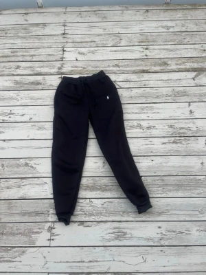 Svarta mjukisbyxor från Ralph Lauren - Säljer ett par svarta mjukisbyxor från Ralph Lauren med dragsko i midjan och resår vid bensluten. Byxorna har två sidofickor och en liten vit Polo-logga broderad på vänster sida. Perfekta för chill dagar eller när du vill vara extra bekväm.