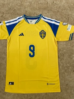 Sverige matchtröja Adidas gul/blå - Gul matchtröja från Adidas för Sveriges landslag med blå detaljer, nummer 9 och namnet ISAK på ryggen. Tröjan har korta ärmar, blåa ränder på axlarna och landslagets emblem på bröstet. Tillverkad i lätt och ventilerande material med Aeroready-teknologi.