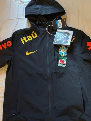 Svart Brasil Nike vindjacka GORE-TEX - Snygg svart vindjacka från Nike med Brasilien CBF-logga, gula och orangea tryck från sponsorer som Itaú och Vivo. Jackan har huva, dragkedja och är tillverkad i GORE-TEX-material som står emot regn och vind. Perfekt för dig som gillar sportig stil och vill sticka ut.