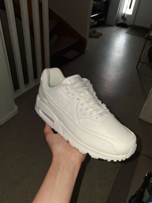 Nike Air Max 90 vita sneakers - Fräscha helvita Nike Air Max 90 sneakers med klassisk chunky sula och synlig Air-enhet i hälen. Skorna har en stilren design i läder och mesh, med vita skosnören och diskreta detaljer. Perfekta för dig som vill ha en clean och tidlös look.