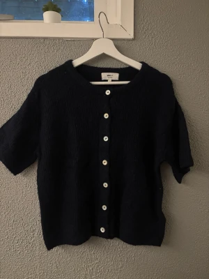 Mörkblå stickad tröja från Gina Tricot - Kortärmad mörkblå stickad tröja från Gina Tricot med rund halsringning och vita knappar framtill. Koftan har en mjuk, lite fluffig struktur och är perfekt för lager på lager. Enkel och stilren design som passar till det mesta.