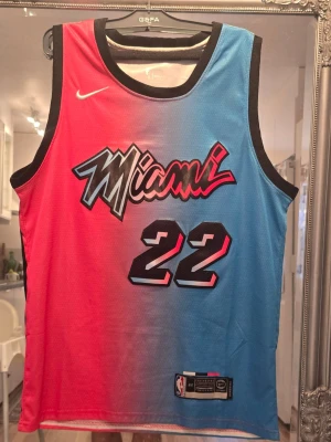 Miami Heat Butler basketlinne #22 - Snyggt basketlinne från Nike med Miami Heat-design i blått och rosa gradient. Nummer 22 och namnet Butler på ryggen. Tillverkad i lätt och ventilerande mesh, perfekt för basket eller streetwear. Svarta kanter runt ärmar och hals för extra kontrast.