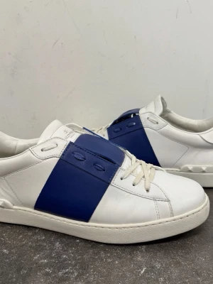 Valentino vita och blå sneakers - Snygga sneakers från Valentino i vitt läder med en bred blå remsa över mitten. Klassisk låg modell med vita snören och diskreta nitar på hälen. Perfekta för dig som gillar stilrena men ändå unika skor.