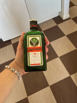 Jägermeister 35cl grön glasflaska - Säljer en klassisk grön glasflaska Jägermeister på 35cl. Flaskan har den ikoniska etiketten med hjorthuvud och orange text. Perfekt till samlingen eller som dekoration. Märket är tydligt präglat på sidan av flaskan. (DEN ÄR HELT SLUT OCH RENGJORD!) mer som dekoration