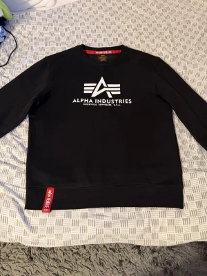 Svart sweatshirt Alpha Industries - Snygg svart sweatshirt från Alpha Industries med vitt tryck och logga på bröstet. Tröjan har rund halsringning, långa ärmar och en röd detalj vid nederkanten. Perfekt för en streetwear-look.