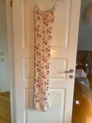 Blommigt nattlinne med smala axelband - Långt nattlinne i ljus färg med rosa och lila blommor. Modellen har smala axelband och en söt knytdetalj vid urringningen. Tyget känns mjukt och luftigt, perfekt för att chilla hemma eller sova bekvämt i.